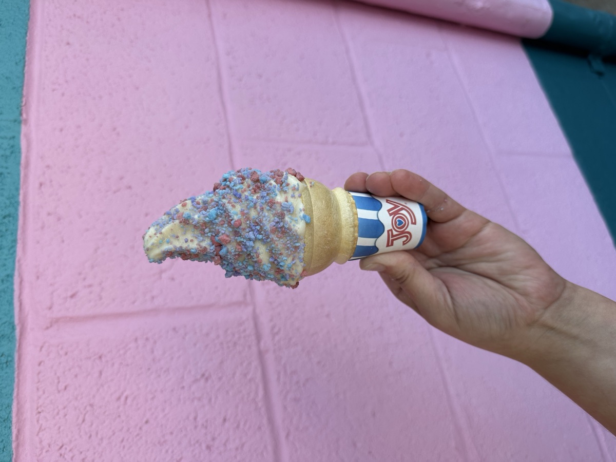Cotton Candy Crunch — a soft serve cone rolled in pink and blue cotton candy crunch, against the pink wall